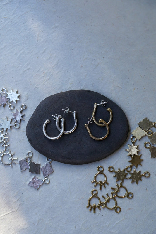 A pair of handmade hoop earrings, one in silver and one in textured bronze, displayed on a dark slate stone. The stone is set against a textured light gray background with different style charms scattered around.