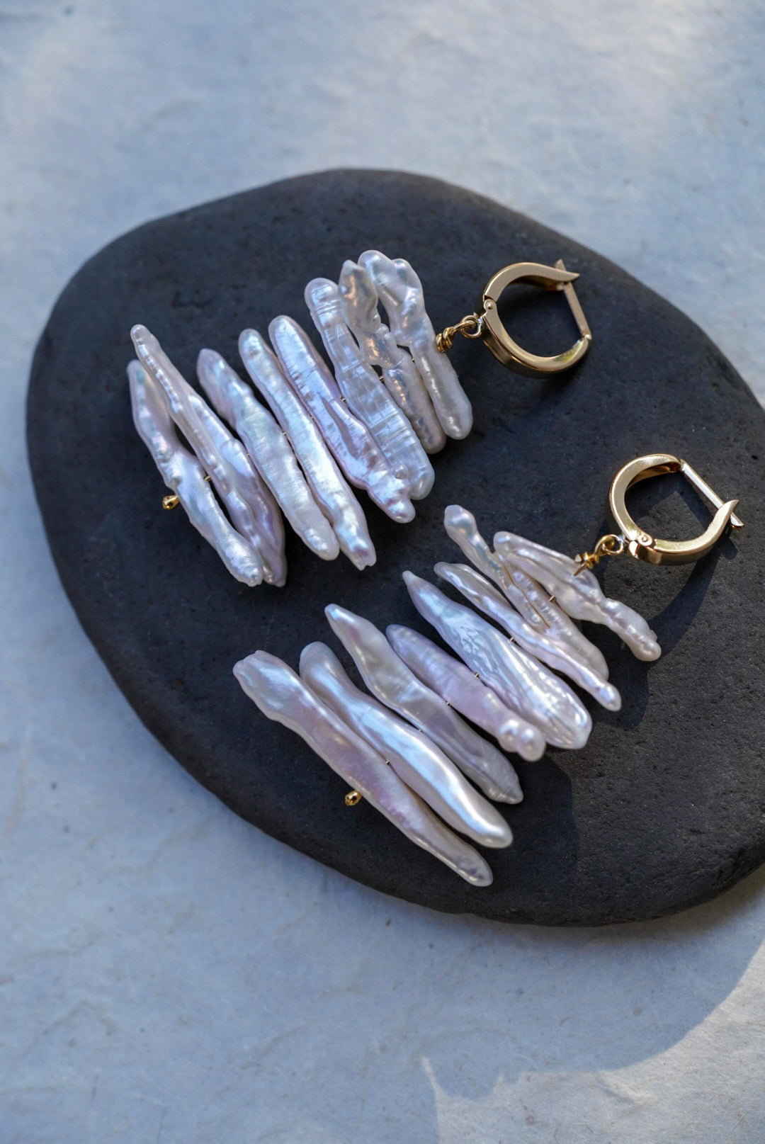 A pair of handmade dangle earrings with gold hoops and stacked iridescent white pearls, displayed on a dark stone against a textured light gray background.