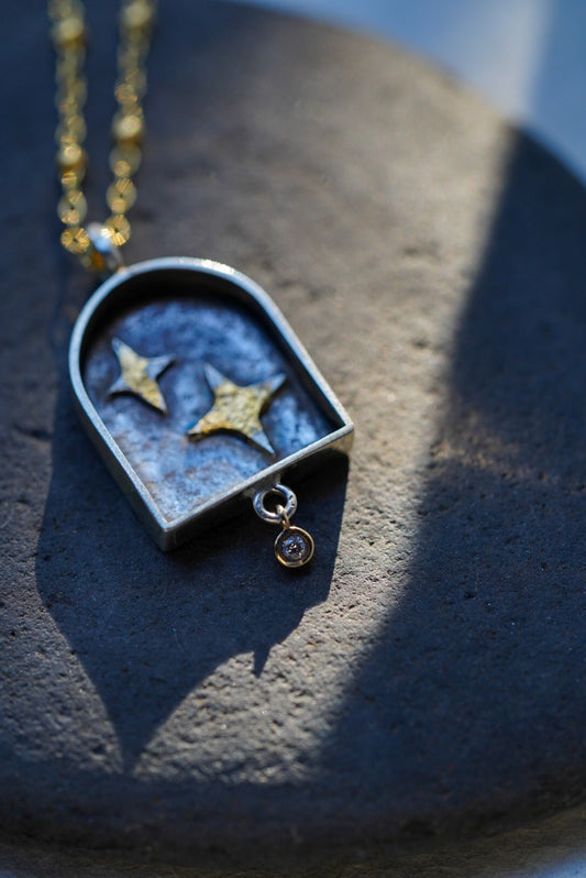A close-up of a gold chain necklace with a arched pendant containing two silver stars with a dusting of gold, resting on a dark stone against a textured light background.