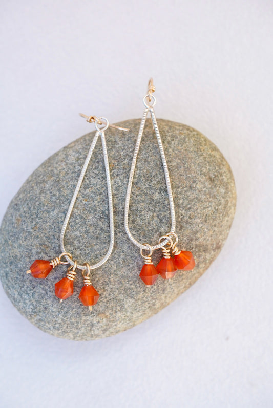 A pair of silver dangle earrings with small orange carnelian bead drops, displayed on a smooth gray stone against a light off-white background.