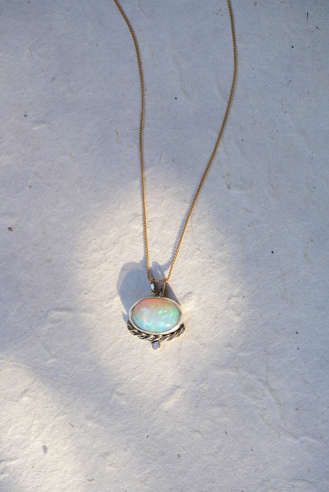 A gold necklace with an oval iridescent opal pendant with a twisted gold accent and small diamond below, against a textured off-white background.