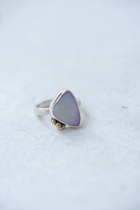 A silver ring featuring a triangular iridescent opal stone with a soft purple and blue sheen, accented by three small gold accents on the stone, set against a light gray textured background.