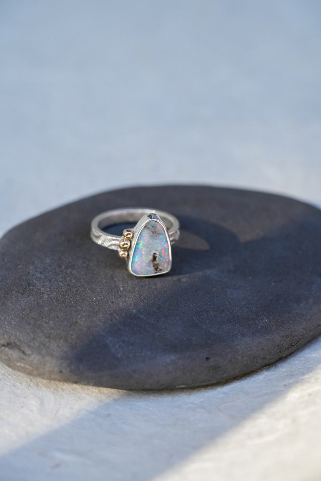 A silver ring with a triangular iridescent opal gemstone and small gold decorative accents on the band, resting on a dark textured surface against a light gray background.