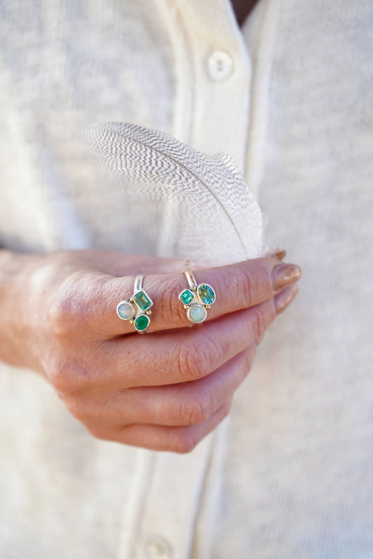 A close-up of a person's hand holding a delicate white feather, wearing two rings with green and pale blue stones, against a soft cream-colored linen shirt.