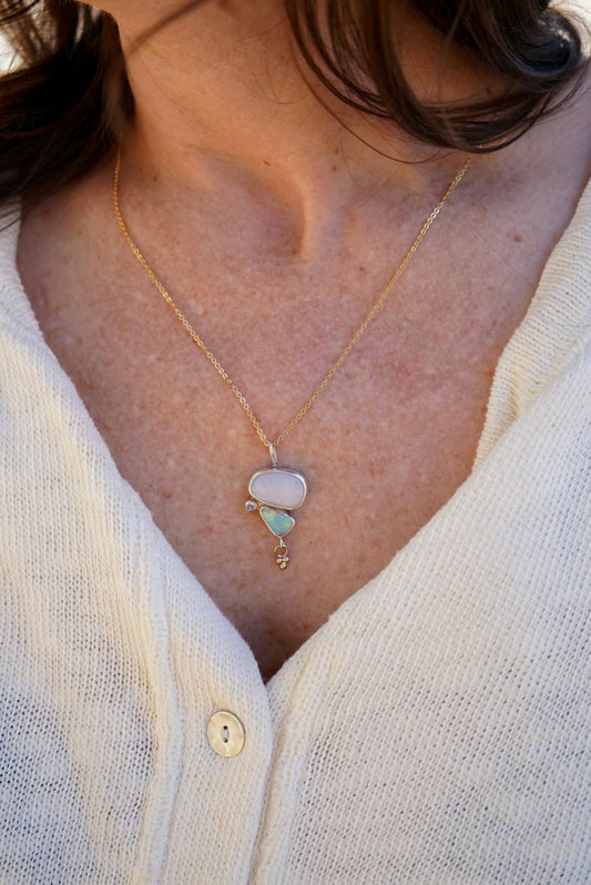A close-up of a person wearing a delicate gold necklace with a pendant featuring a pale pink opal stone, a mint green opal stone, and a tiny gold charm, paired with an off-white knit top.