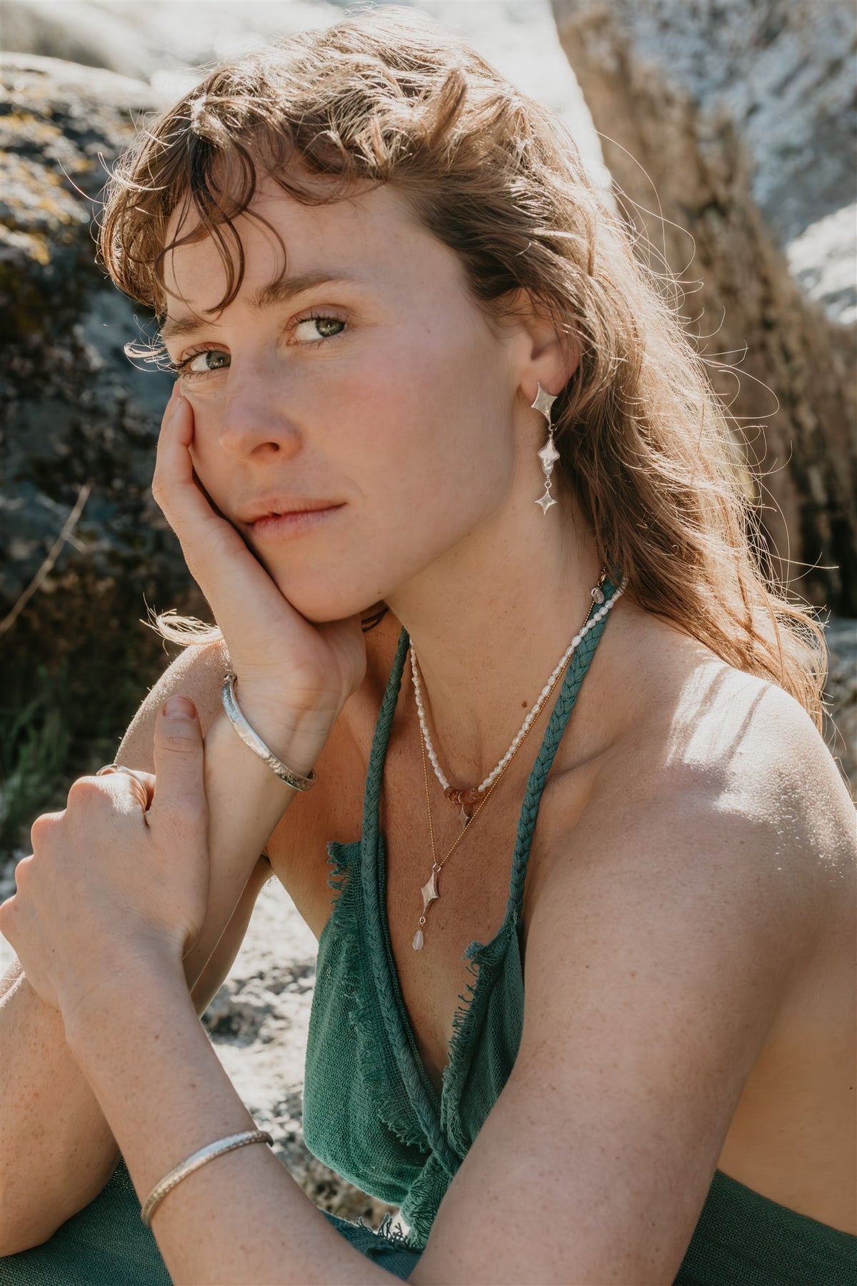 Woman with long hair and green top sitting outdoors near rocks, wearing earrings, bracelets, and necklaces