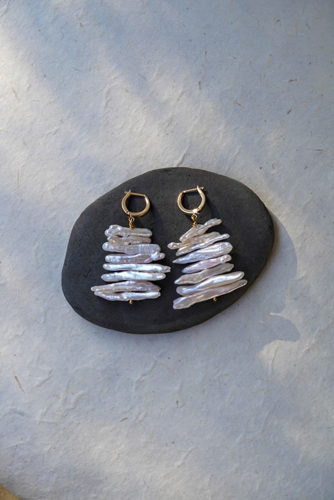 A pair of handmade dangle earrings with gold hoops and stacked iridescent white pearls, displayed on a dark stone against a textured light gray background.