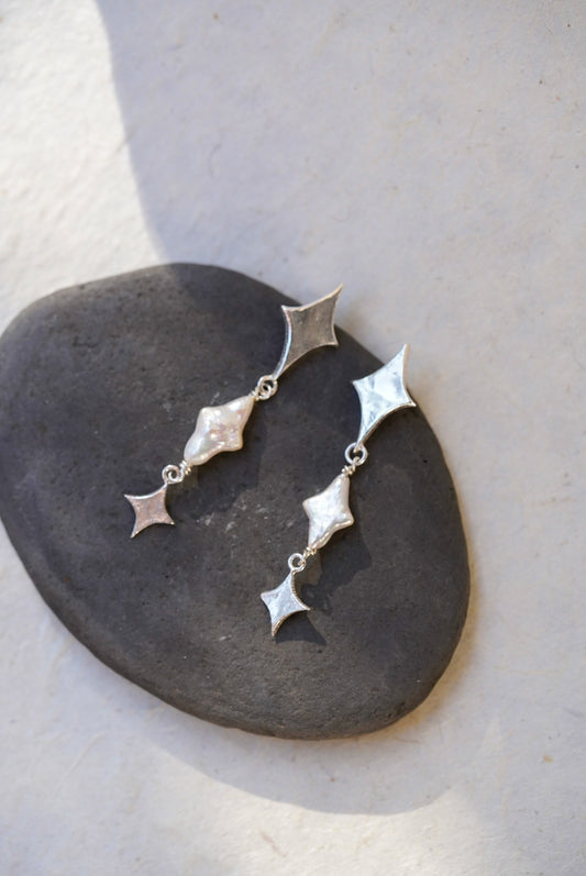A pair of silver dangle earrings with star-shaped and star pearls, displayed on a dark stone against a light beige background.