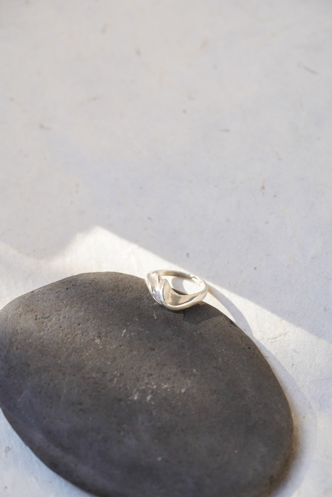 A polished silver ring with a curved, organic shape resting on a smooth dark gray stone against a light gray background.