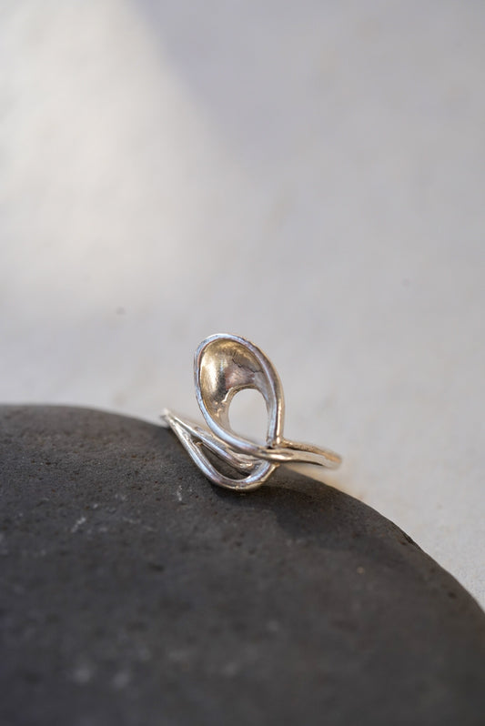 A silver abstract curved ring with a polished finish, resting on a dark textured stone against a light gray background.