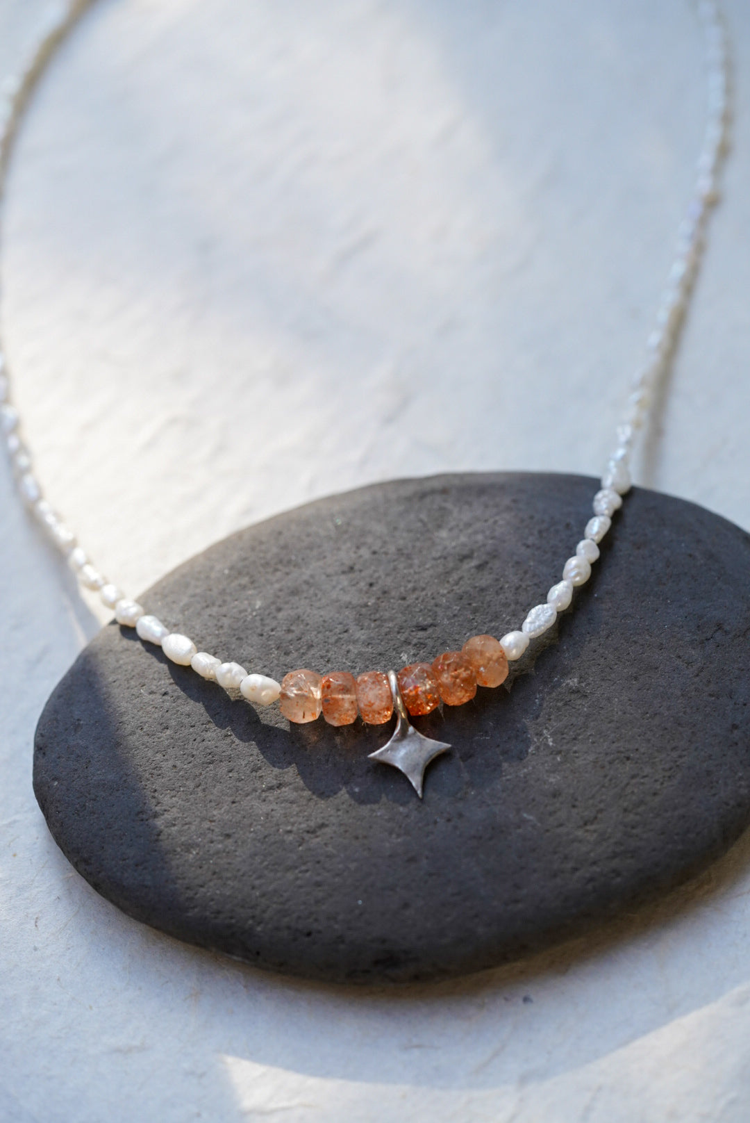 A delicate beaded necklace with white pearl beads, a row of faceted sunstone stones, and a small silver star charm, displayed on a dark stone over a light textured surface.
