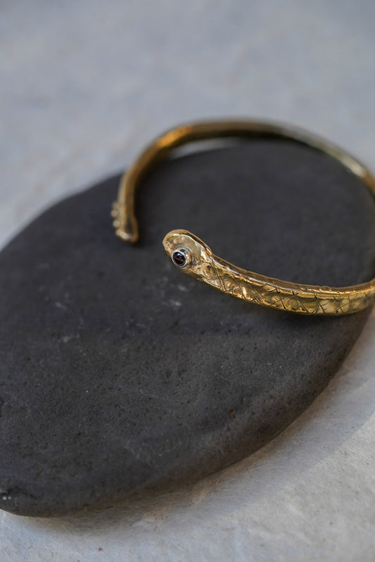 A bronze snake cuff with a small garnet, resting on a dark stone against a light gray background.