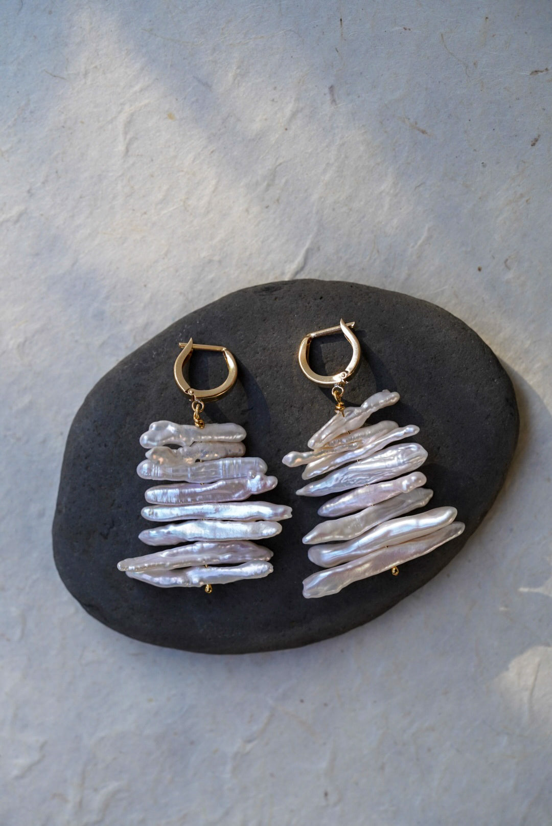 A pair of handmade dangle earrings with gold hoops and stacked iridescent white pearls, displayed on a dark stone against a textured light gray background.