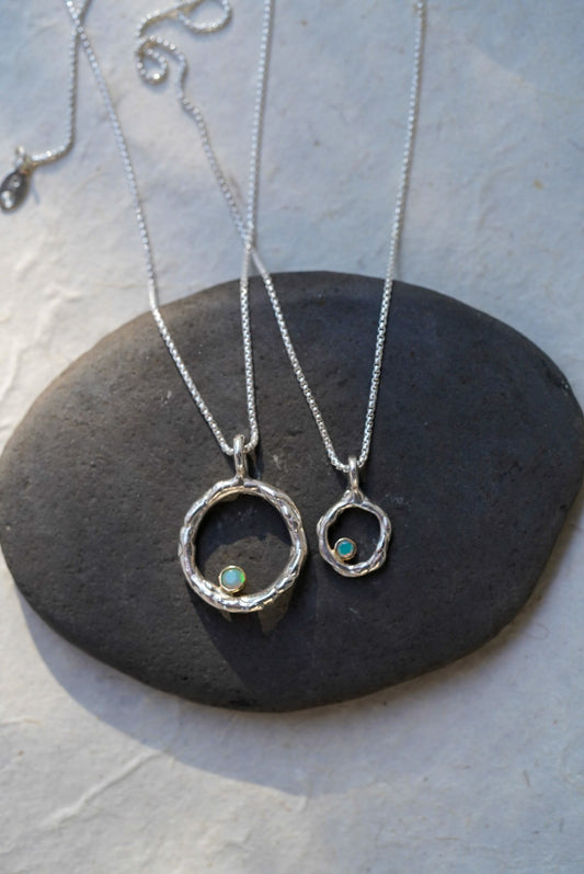Two silver chain necklaces with open circular pendants, each featuring a small iridescent opal stones, displayed on a dark stone slab against a light textured background.