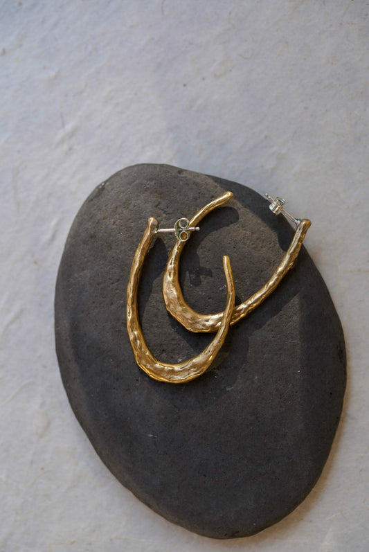 A pair of textured gold hoop earrings displayed on a dark gray stone against a light textured background.