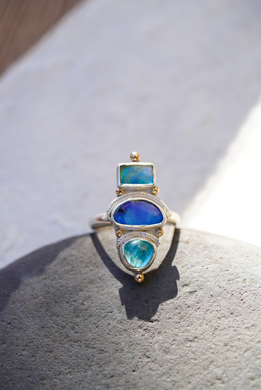 A silver ring with three stacked bright blue stones in vibrant blue and turquoise tones, displayed on a piece of rock  against a neutral light background.