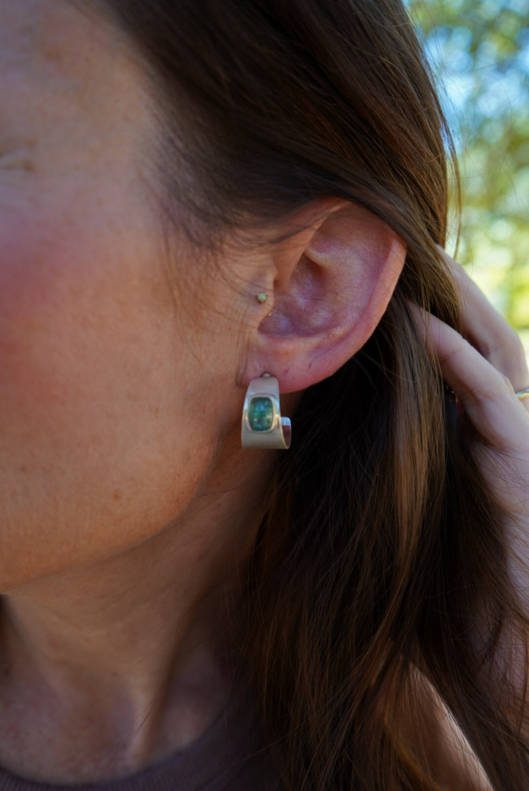 A close-up of a person's ear wearing a small silver hoop earring with a green gemstone, against an outdoor blurred background.