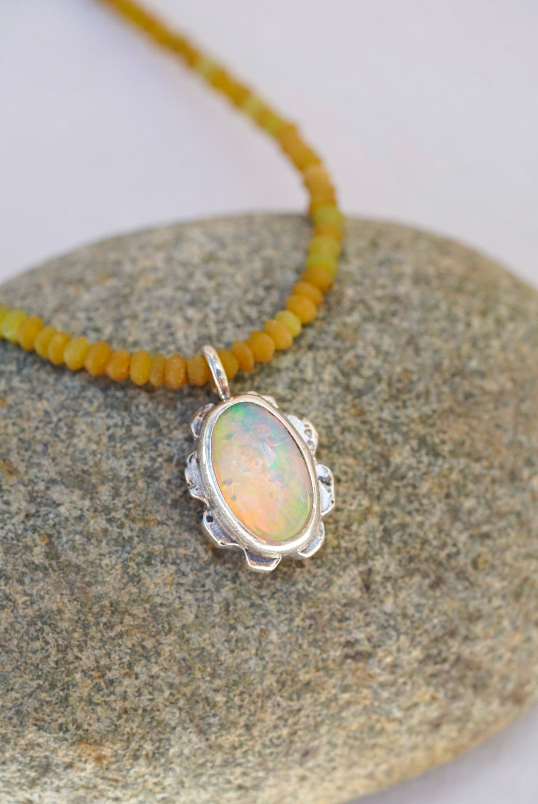 A silver pendant necklace with an oval iridescent opal stone, resting on a smooth gray stone with a yellow beaded strand visible in the background.