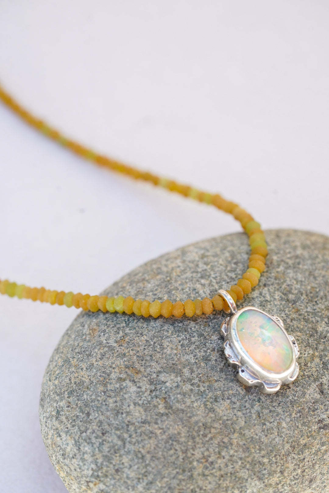 A close-up of a necklace with a silver-toned oval pendant featuring an iridescent opal-like stone, hanging over a smooth gray stone on a light background.