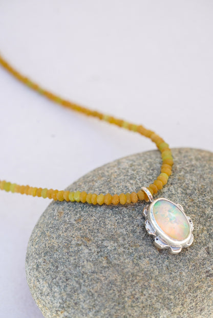 A close-up of a necklace with a silver-toned oval pendant featuring an iridescent opal-like stone, hanging over a smooth gray stone on a light background.