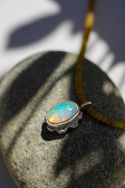 A silver pendant necklace with an oval iridescent opal gemstone resting on a textured gray stone, with a yellow beaded necklace strand visible in the background under dappled sunlight.