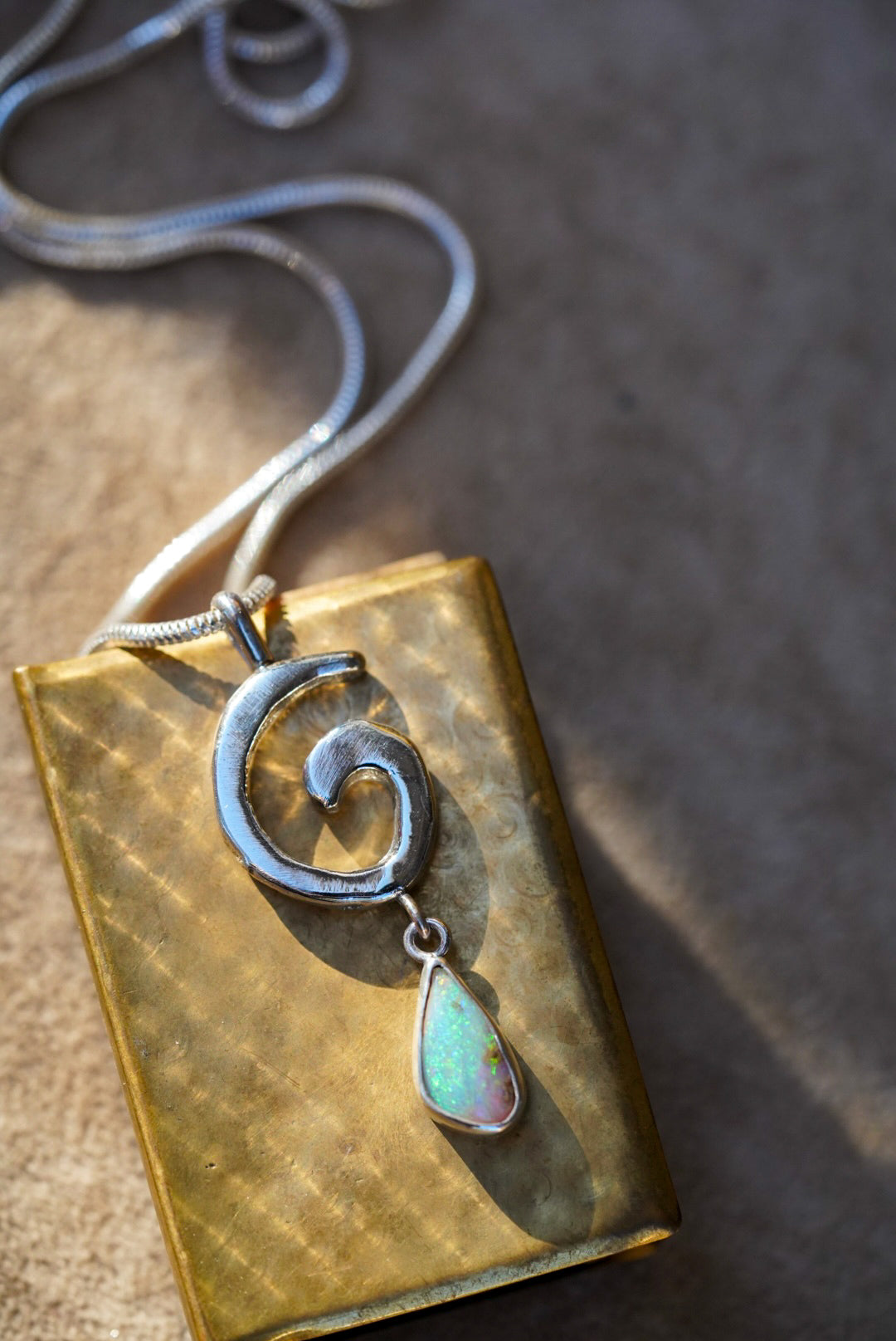 A silver necklace with a spiral pendant and a teardrop-shaped iridescent opal drop, displayed on a dark stone against a vintage gold matchbox on a textured off-white background.