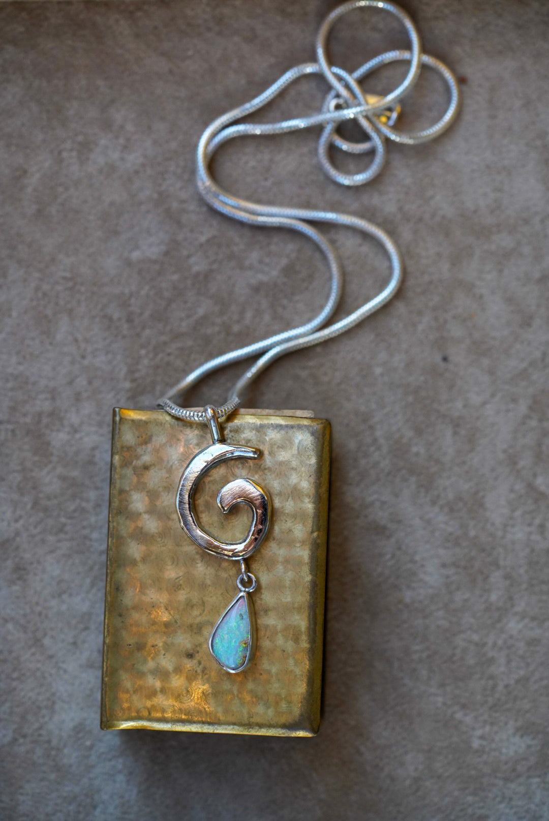 A silver necklace with a spiral pendant and a teardrop-shaped iridescent opal drop, displayed on a dark stone against a vintage gold matchbox on a textured off-white background.