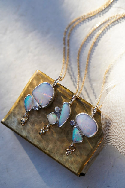 Three silver necklaces featuring opals and a moonstone along with a small gold charm below, displayed on a textured vintage gold matchbox against a light neutral background.