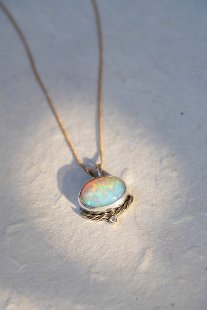 A gold necklace with an oval iridescent opal pendant with a twisted gold accent and small diamond below, resting on a dark gray stone against a textured off-white background.