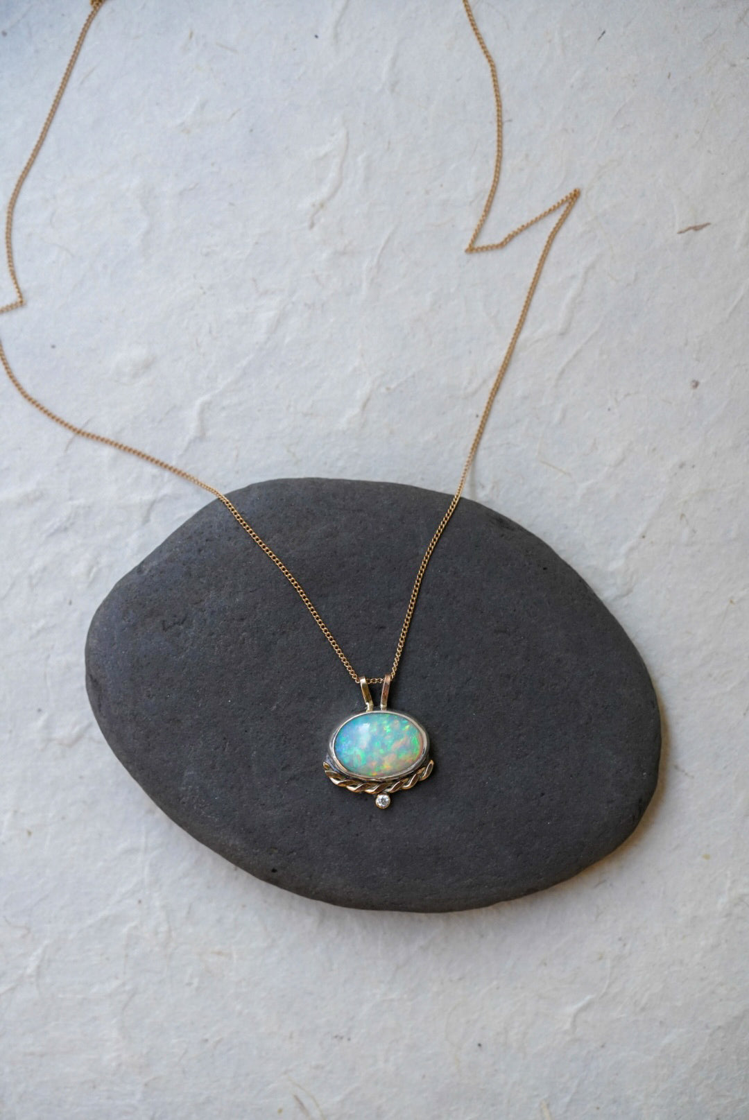 A gold necklace with an oval iridescent opal pendant with a twisted gold accent and small diamond below, resting on a dark gray stone against a textured off-white background.