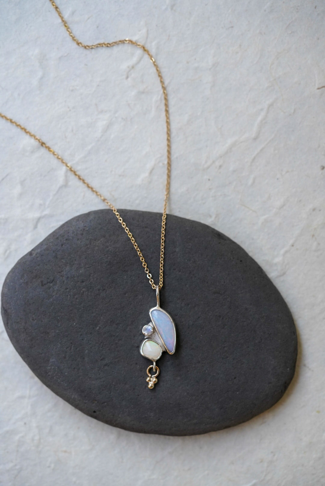 A delicate gold chain necklace with a pendant featuring a curved iridescent opal stone, another small opal stone, and a moonstone, and a tiny dangling gold charm, displayed on a dark gray stone against a light textured background.