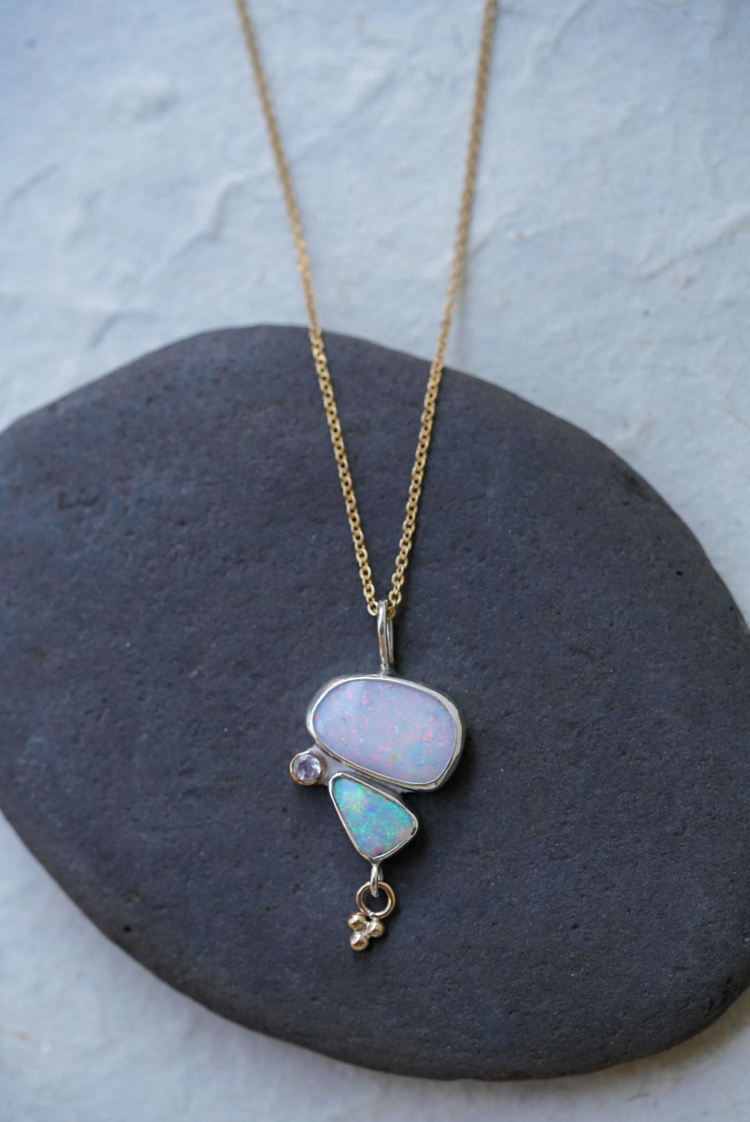 A gold chain necklace with a unique pendant featuring a large opal stone and a smaller light blue-green opal stone alongside a moonstone and a small charm underneath. Displayed on a dark gray stone against a textured off-white background.