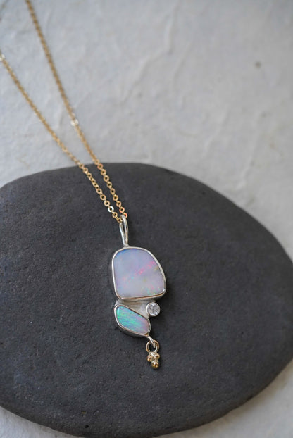 A gold chain necklace with a silver pendant featuring a large iridescent opal and a smaller green-blue opal opal, accented by a moonstone and a dangling gold bead, displayed on a dark gray stone against a light textured background.