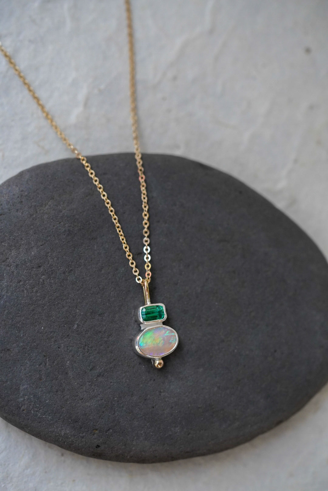 A delicate gold chain necklace with a silver pendant featuring a green rectangular tourmaline and an oval iridescent opal, displayed on a dark gray stone against a light gray background.