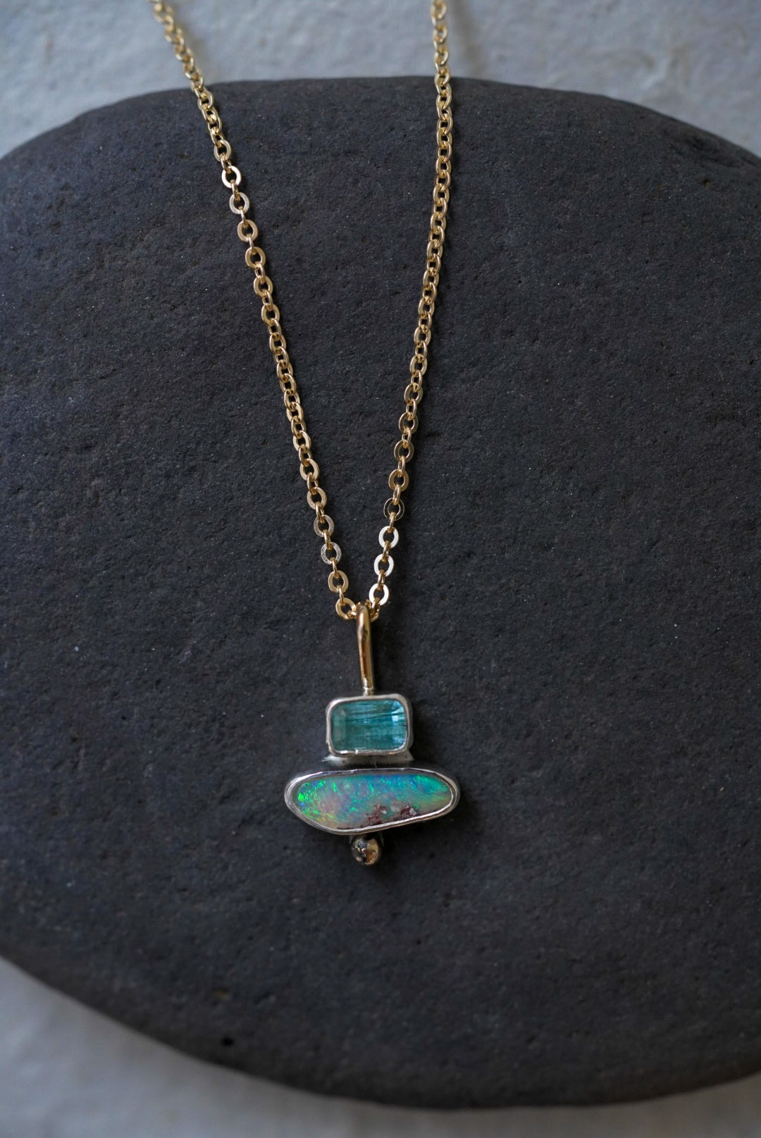 A delicate gold chain necklace with a pendant made of stacked natural stones, one tourmaline and one opal, displayed on a dark gray stone slab against a textured off-white background.