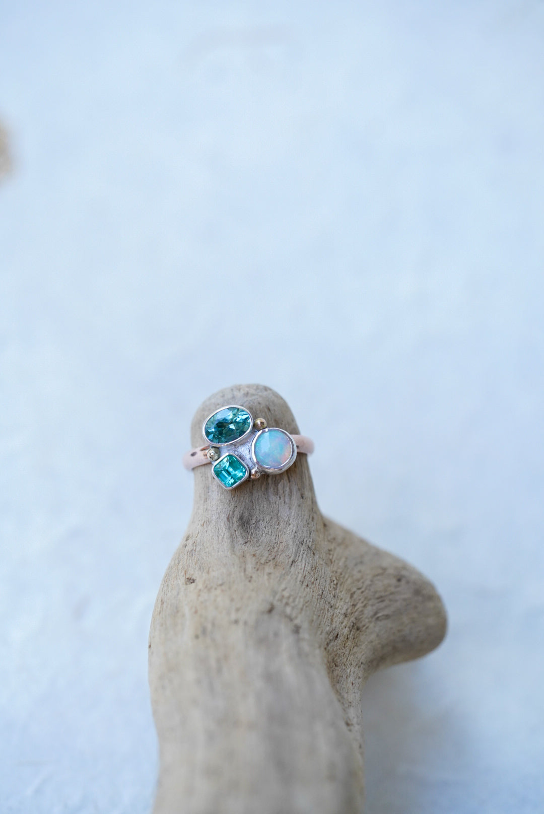 A rose gold ring with three green-blue gemstones, resting on a pice of driftwood against a light gray background.