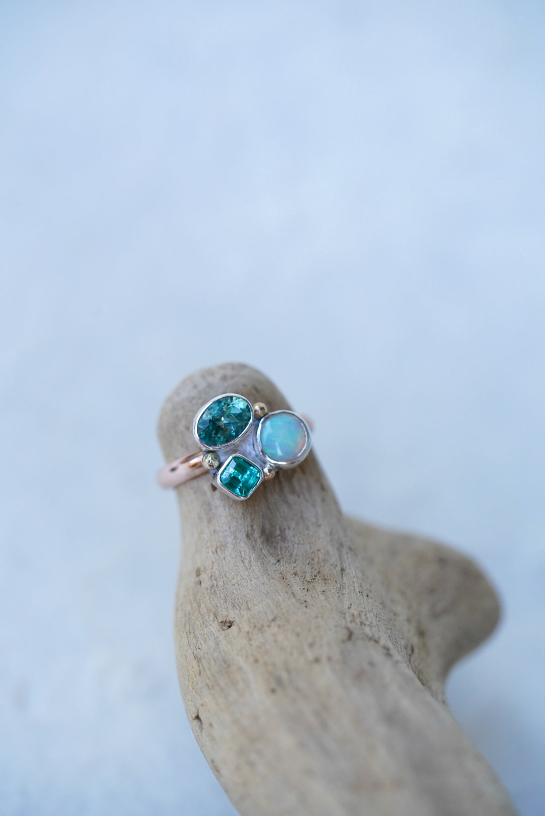 A rose gold ring with three green-blue gemstones, resting on a pice of driftwood against a light gray background.