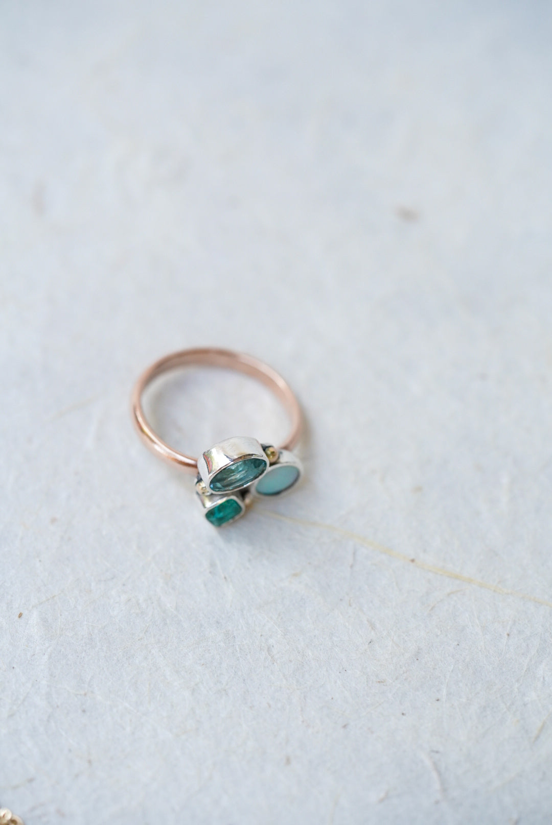 A delicate ring with a rose gold band and a cluster of gemstones in a silver setting, resting on a textured light gray surface.
