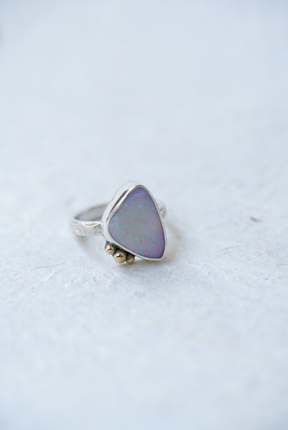 A silver ring featuring a triangular iridescent opal stone with a soft purple and blue sheen, accented by three small gold accents on the stone, set against a light gray textured background.