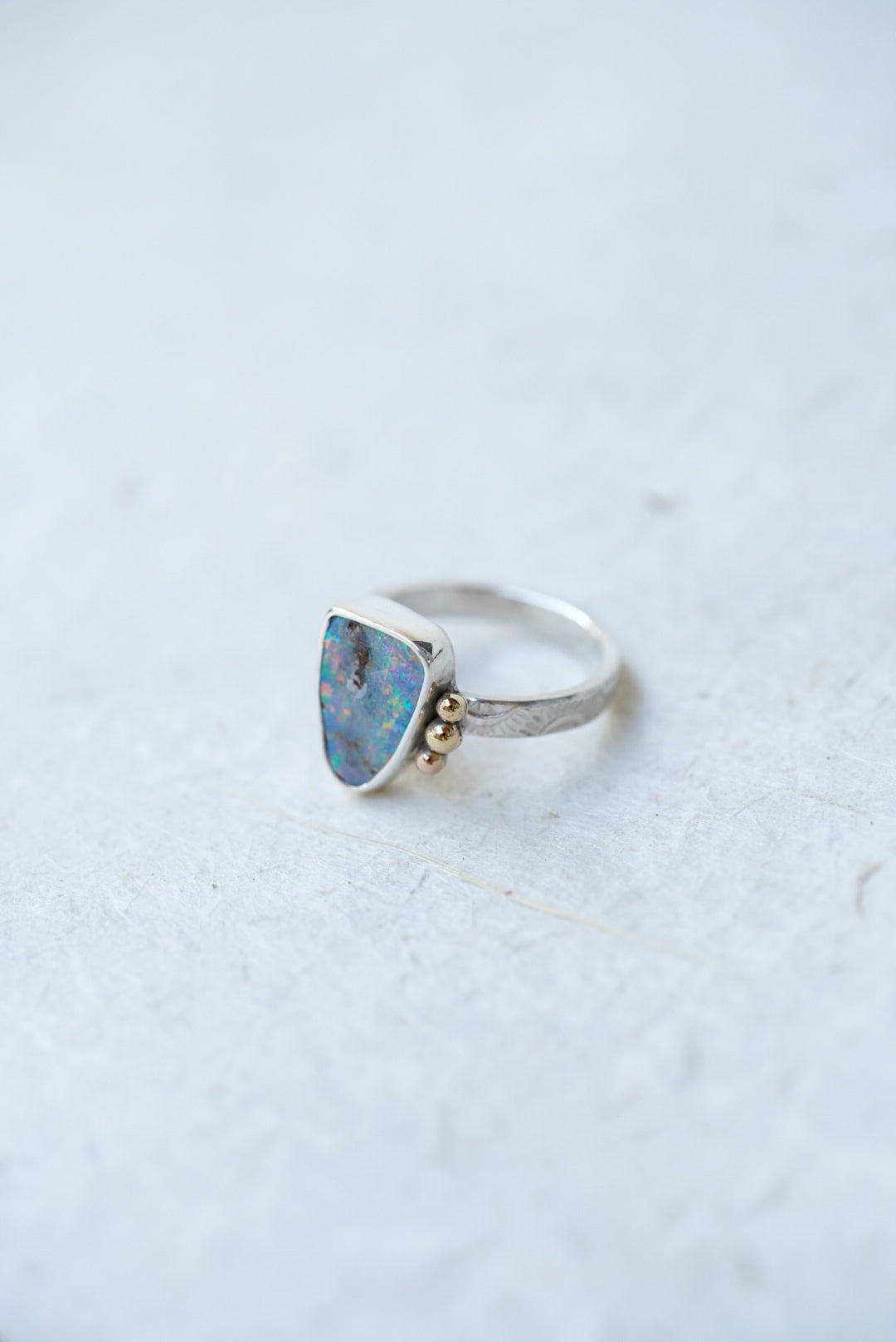 A silver ring featuring a triangular opal stone in a bezel setting, accented with small gold accents on the stone, set against a light textured background.