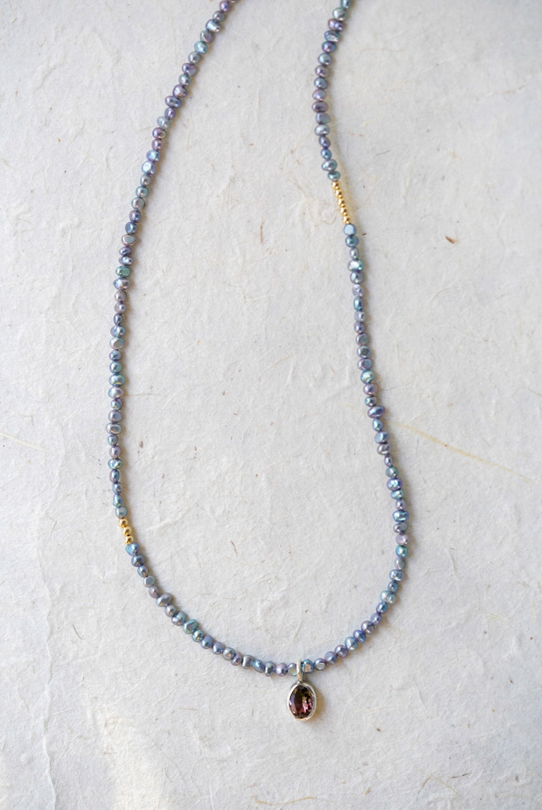 A delicate beaded necklace with small iridescent blue-gray pearls, gold spacer accents, and a tourmaline at the center, laid on a light textured surface.