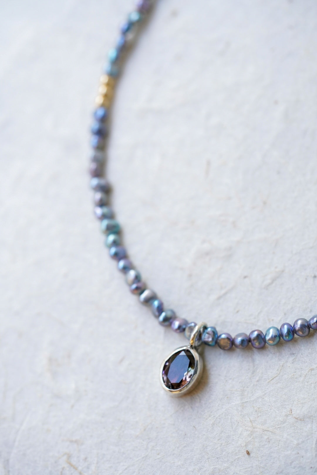 A closeup of a delicate beaded necklace with small iridescent blue-gray pearls, gold spacer accents, and a tourmaline at the center, laid on a light textured surface.