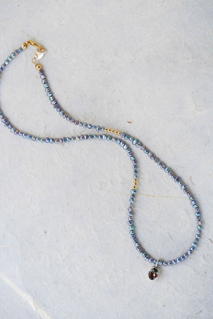 A delicate beaded necklace with small iridescent blue-gray pearls, gold spacer accents, and a tourmaline at the center, laid on a light textured surface.