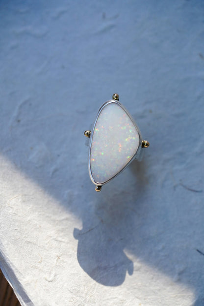 A triangular bezel-set ring featuring a white opal stone with iridescent flashes, held in a silver setting with small gold-toned accents, resting on a textured light surface.