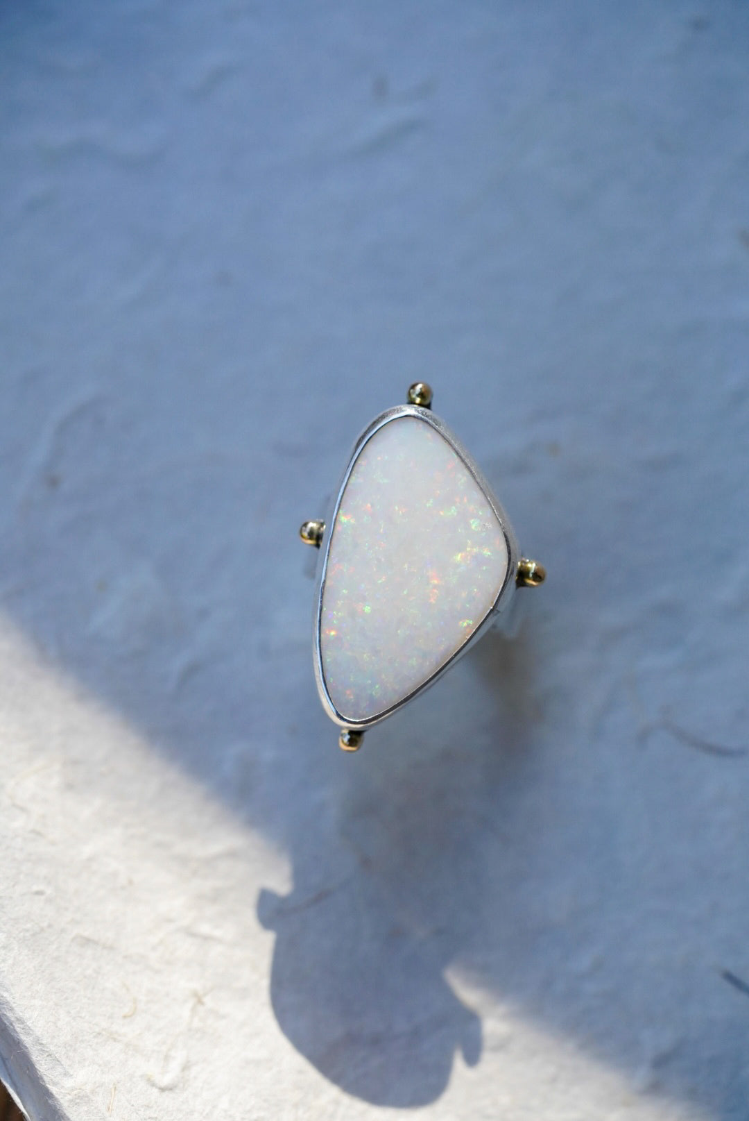 A triangular bezel-set ring featuring a white opal stone with iridescent flashes, held in a silver setting with small gold-toned accents, resting on a textured light surface.