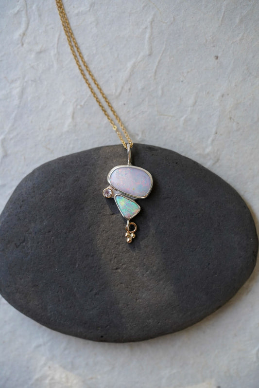A gold chain necklace with a unique pendant featuring a large opal stone and a smaller light blue-green opal stone alongside a moonstone and a small charm underneath. Displayed on a dark gray stone against a textured off-white background.