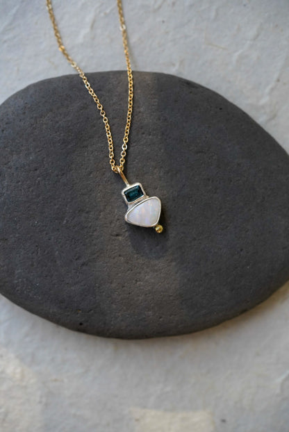 A delicate gold chain necklace with a small silver pendant featuring a white teardrop-shaped opal stone, a tiny dark tourmaline square, and a small gold bead, displayed on a dark gray stone against a light gray background.