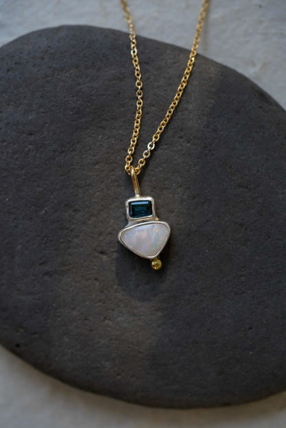 A delicate gold chain necklace with a small silver pendant featuring a white teardrop-shaped opal stone, a tiny dark tourmaline square, and a small gold bead, displayed on a dark gray stone against a light gray background.