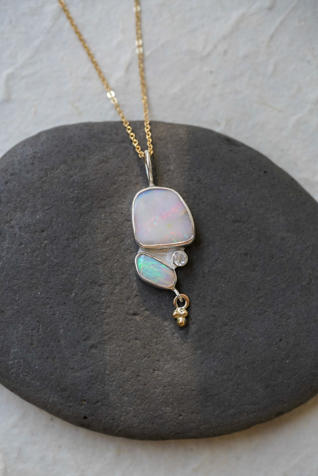 A gold chain necklace with a silver pendant featuring a large iridescent opal and a smaller green-blue opal opal, accented by a moonstone and a dangling gold bead, displayed on a dark gray stone against a light textured background.