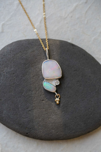 A gold chain necklace with a silver pendant featuring a large iridescent opal and a smaller green-blue opal opal, accented by a moonstone and a dangling gold bead, displayed on a dark gray stone against a light textured background.